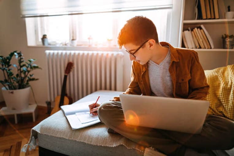 young man with glasses doing homework at his desk by his bedroom window