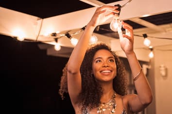 Cheerful young woman wearing party dress changing bulb light in patio. Happy smiling girl making preparation of party by adding lights outdoor. 