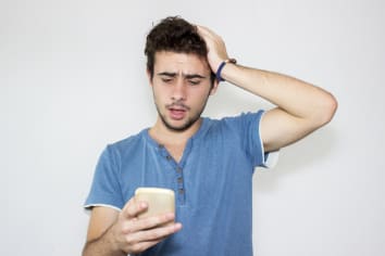 Surprised young man in blue t-shirt standing in front of gray background reading a message with frustrated facial expression. 