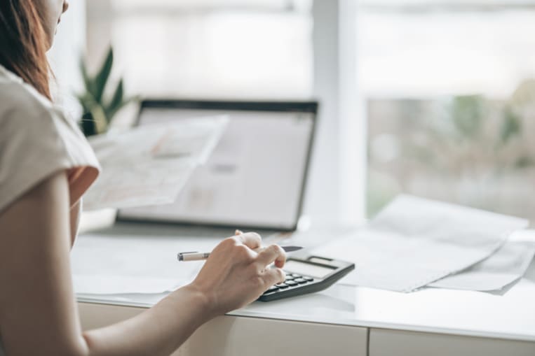 Woman in white shirt sits in front of a computer. Women is typing on a calculator while looking at a computer screen. 