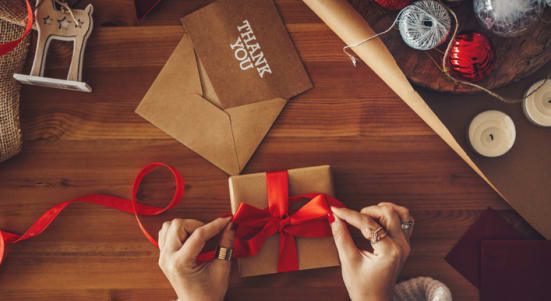Hands in a white sweater tie a bow on a present. On the table nearby, sits is a card reading "thank you"