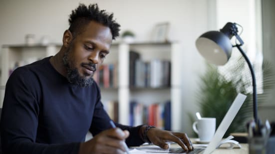Man working at home. Man sits at desk and uses laptop while making notes on a sheet of paper. 