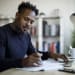 Man working at home. Man sits at desk and uses laptop while making notes on a sheet of paper. 