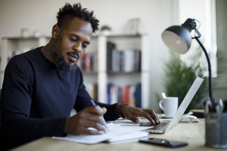 Man working at home. Man sits at desk and uses laptop while making notes on a sheet of paper.