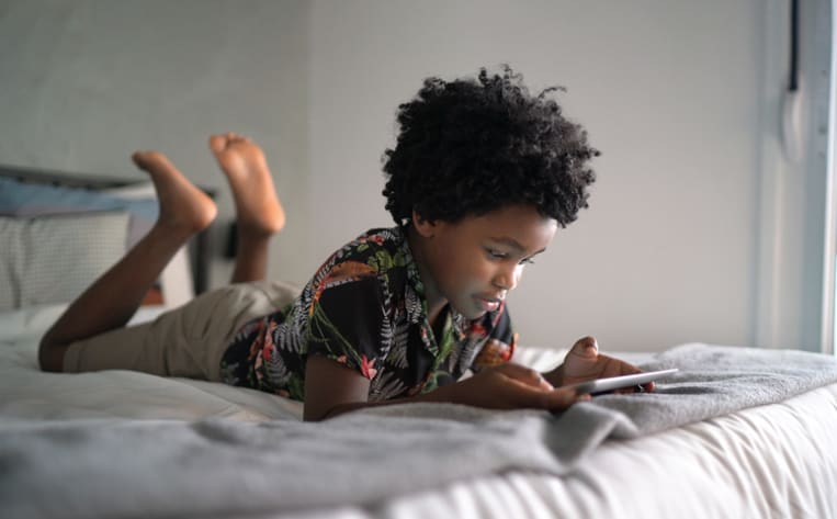 A boy about 5 years old lays on a bed playing with a tablet or mobile phone.