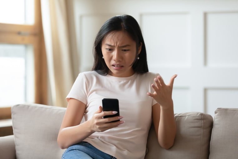 Young woman sits on couch and looks in anger and frustration at her phone. 