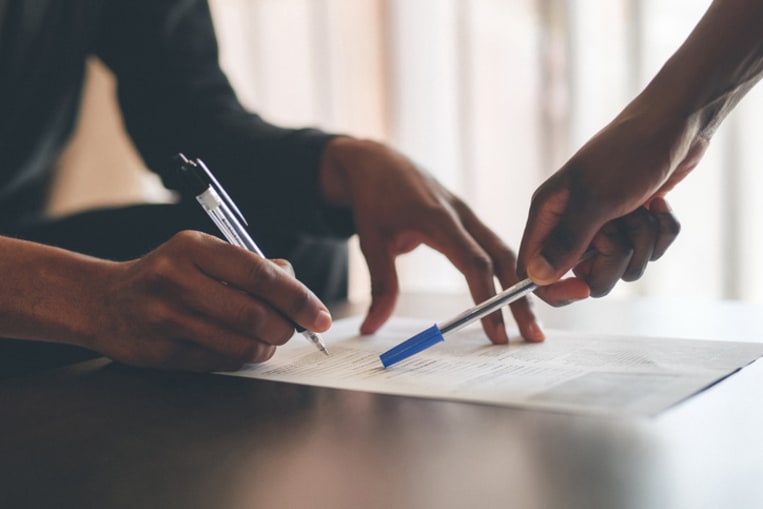 A hand points out where to sign a contract, while another person holding a pen signs.