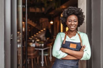 Black woman in an apron standing in front of a business smiling