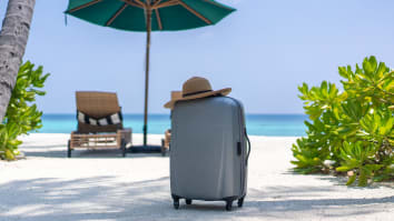 A suitcase sits on the beach near a beach chair and umbrella. A sun hat is balanced on top of the suitcase.