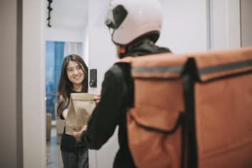 Man wearing a bike helmet and backpack delivers food to a young woman at an apartment door. 