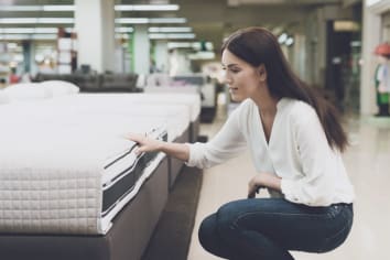 A woman in a white shirt and jeans in a mattress store. She examines the mattress she wants to buy. She squats and looks at the mattress�