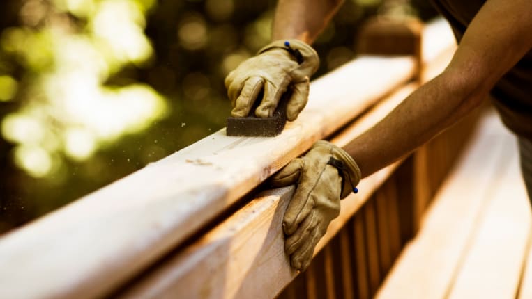 A close-up on a contractor's gloved hands sanding a wooden deck rail.