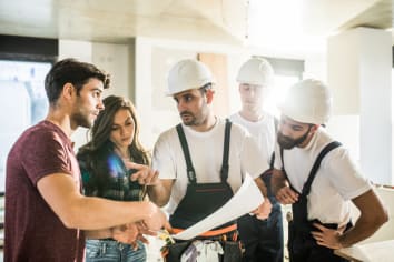 Young woman and man viewing building plans with three male contractors in white hard hats and black overalls