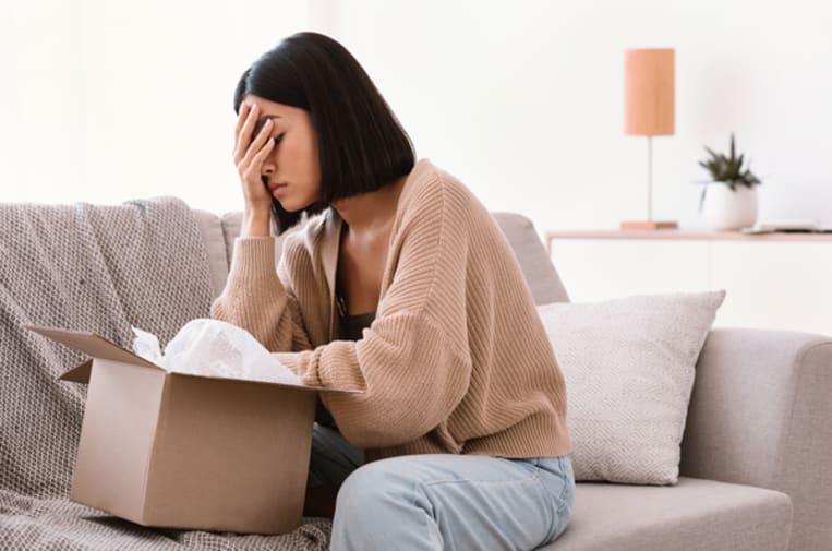 Sad upset Asian lady sitting on the couch and unpacking a cardboard box.