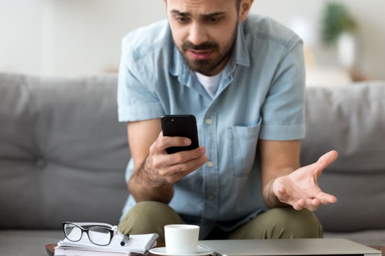 Close up confused man having problem with broken not working phone, annoyed frustrated male student receiving bad news, reading unpleasant email, sitting on sofa, looking at screen, spam message