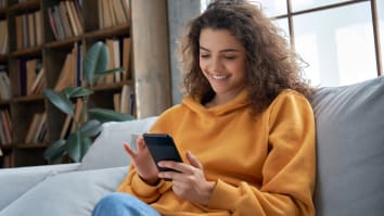 A young woman wearing an orange sweatshirt sits on a couch. She is smiling and looking at her cell phone.