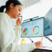 A woman in front of two monitors looks at data reports and writes in a notebook. 