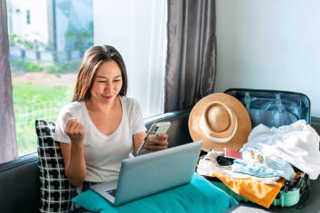 Young woman sits in front of a packed suitcase looking at her laptop and smartphone. She is smiling and the suitcase overflows with vacation clothes, such a sun hat. 