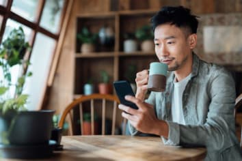 Young Asian man sits at a kitchen table. He looks at his cell phone while drinking from a mug. 