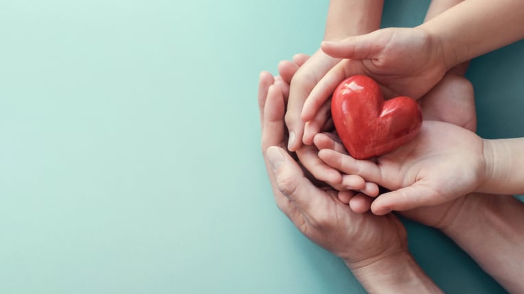 Three pairs of hands, both adults and children, hold a small red ceramic heart.