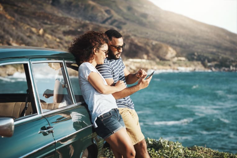 A young couple leans against a car overlooking a body of water. They are looking a call phone. 