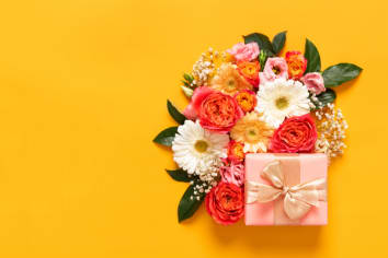 Orange, white, and yellow-colored flowers bouquet fixated around a pink wrapped present. The flowers are displayed off-center over an orange background.