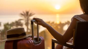 A tourist woman is sitting with a suitcase and straw hat and looking at an ocean sunset.