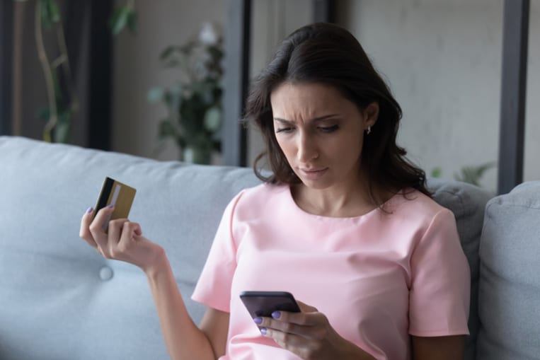 Unhappy woman holds a credit card in one had and a smartphone in another while sitting on the couch. 