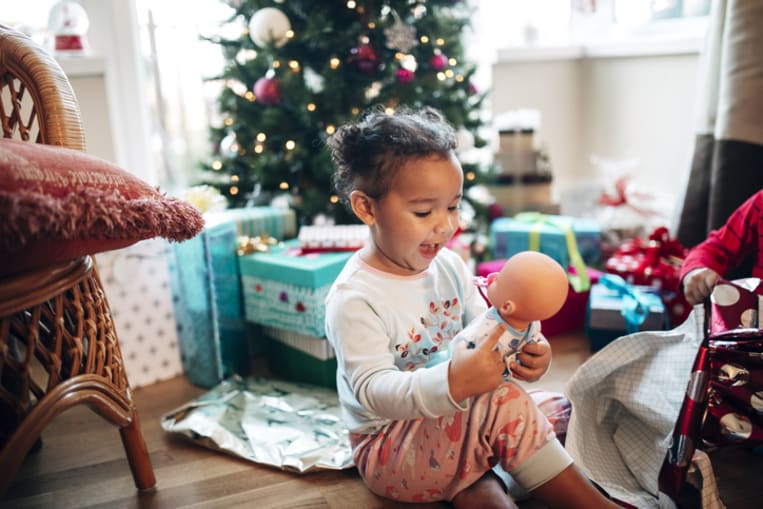 Toddler girl sits on floor next to a Christmas tree and happily opens the present of a baby doll 