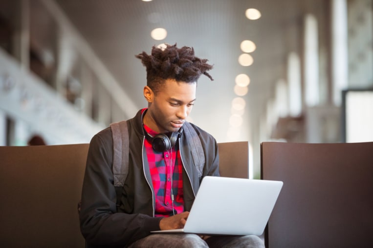 Young man, dressed casually using laptop while sitting on seat waiting at airport.