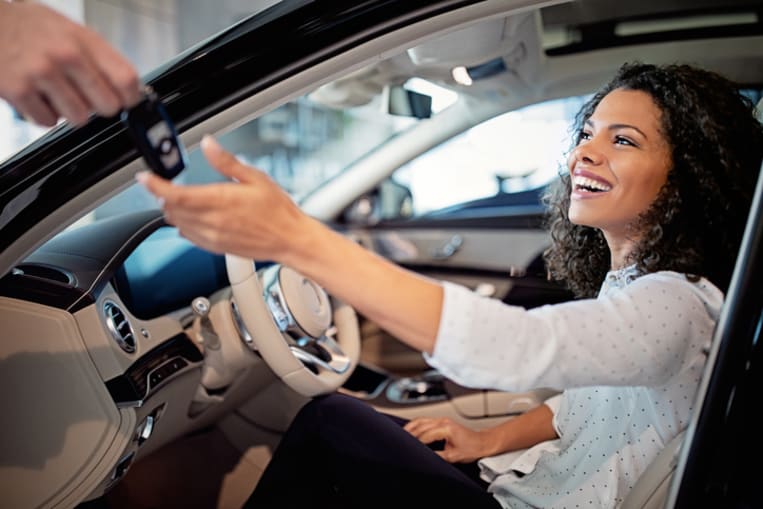 Someone outside the photo hands a smiling woman, who sits behind the steering wheel of a new car, a set of car keys. 