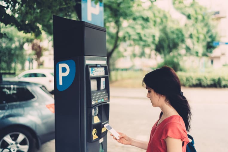 Woman with long dark hair, wearing a pink tshirt stands in a parking lot and pays with her mobile phone