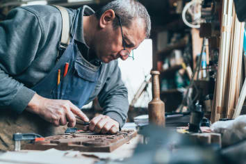 A man with glasses and gray hair leans over his workbench and carves into a wooden block. 