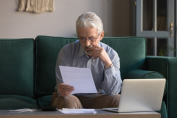 Serious mature man wearing glasses sitting on couch in living room manage budget received invoice analyzes month expenses feels concerned about public utility debt, check read loan documents concept