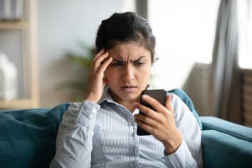 Confused millennial Indian girl stares at phone in frustration with a hand on her forehead