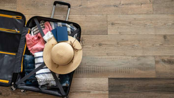 An open suitcase rests on a wooden floor. It contains clothes, a sun hat and a passport.