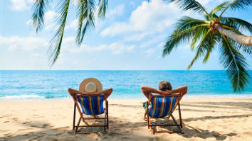 Two vacationers recline in beach chairs under palm trees and look at the ocean.