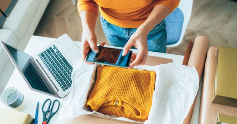Woman taking photo of yellow sweater in shipping box with laptop beside it