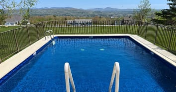 View of pool from top of ladder. Pool is above ground with blue lining, filled with water and a fence around the top. There are views of the mountains of NH.