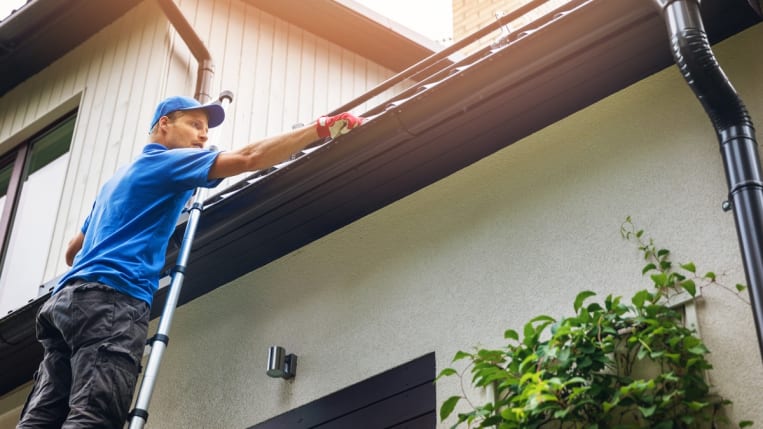 A gutter contractor stands on a ladder to clean the gutter on a home.