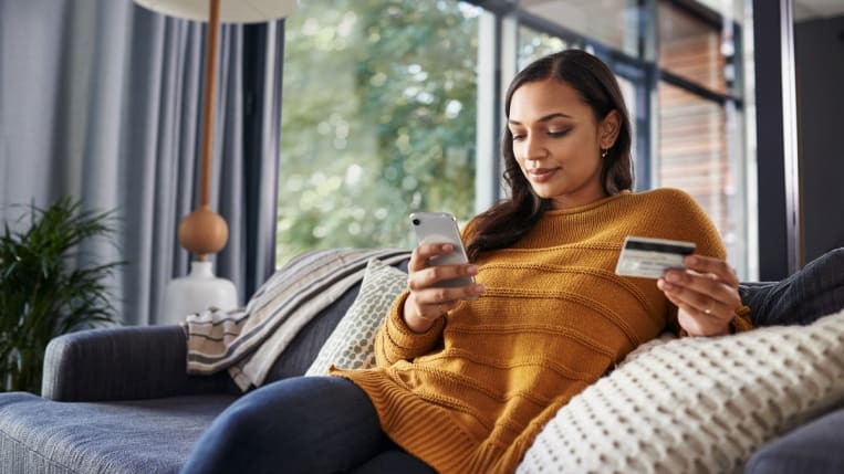 A young woman is sitting on a couch and using a smartphone to shop online.