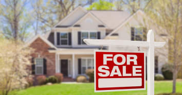 Brick and white siding house blurred in background surrounded by trees with red For Sale sign in front
