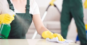 Person wearing green overalls and yellow rubber gloves sprays green cleaner on countertop to clean.