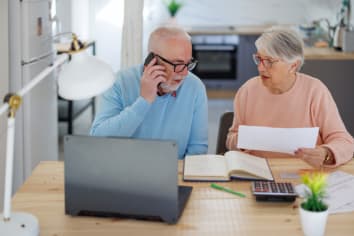 A senior couple uses a laptop to pay their bills while sitting at a table in their house. Frustrated senior couple sitting at home and checking their home finances
