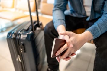 Travel abroad concept. Male tourist hand holding passport sitting with suitcase luggage on seat in waiting area of airport terminal.