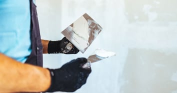 Contractor wearing blue shirt, apron and black gloves applies plaster to drywall