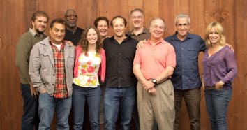 The Such a Voice team of nine people standing against a wood panel wall