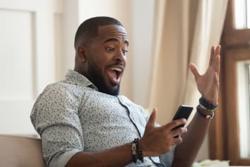 Surprised happy african man holding phone looking at cellphone read good news in sms sit on sofa