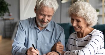 Senior couple smiling while signing paperwork. The woman has her arm linked with the man's arm and is wearing white and black striped shirt. The man is wearing a blue button down shirt.