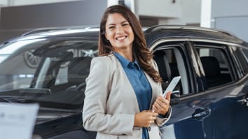 Smiling saleswoman holding tablet PC while looking at camera at new car showroom. Professional car dealer posing proudly at auto showroom, smiling to the camera, holding tablet PC.	
